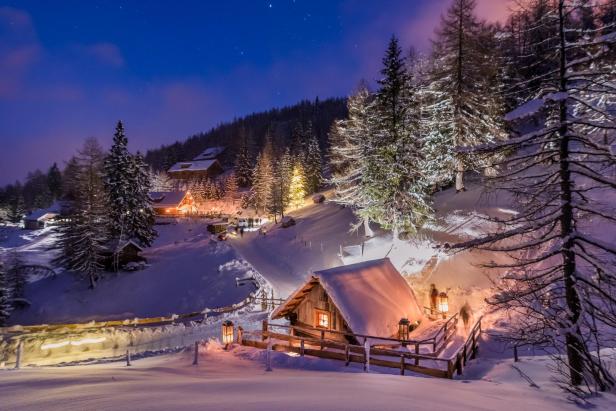 Verschneite Berghütte mit warmem Licht, umgeben von schneebedeckten Bäumen und einem Zaun, Abendstimmung am Katschberg.