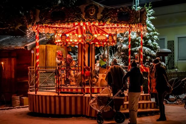 Beleuchtetes Kinderkarussell mit rot-weißen Streifen und Figuren, umgeben von mehreren Personen und Schnee bei Nacht.