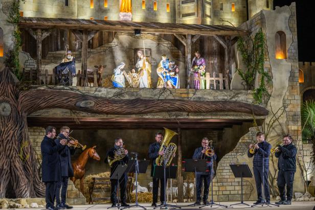 Eine Bläsergruppe spielt vor einer großen, beleuchteten Weihnachtskrippe mit Figuren der Heiligen Familie und den Heiligen Drei Königen.