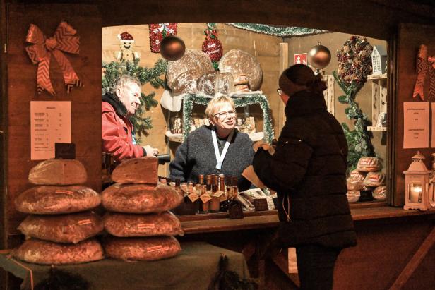 Weihnachtsmarktstand mit drei Personen, die Backwaren verkaufen, und einer Kundin, die etwas auswählt. Der Stand ist mit Tannenzweigen, Kugeln und Schleifen dekoriert.