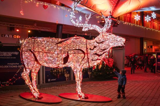Leuchtender Rentier-Dekorationsaufbau aus Lichterketten mit zwei Kindern davor in einer beleuchteten Einkaufsstraße.