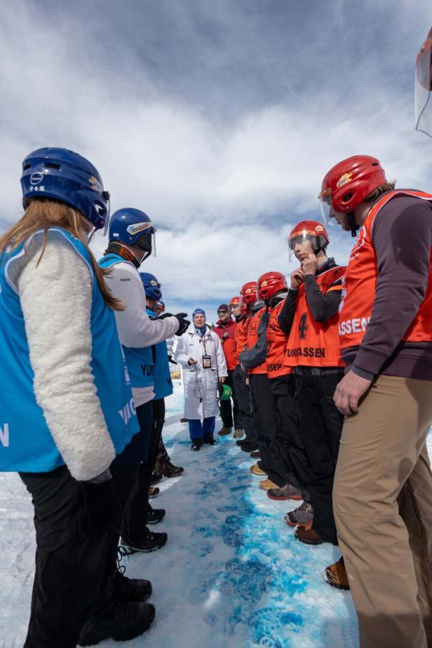Zwei Gruppen von Personen stehen sich auf einem schneebedeckten Weg gegenüber, eine Gruppe trägt blaue Helme und Westen, die andere rote Helme und Westen.