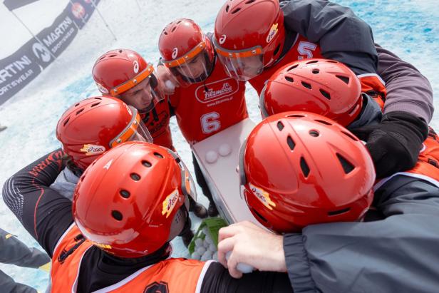 Gruppe von Personen mit roten Helmen und roten Westen, die sich im Kreis zusammenbeugen, im Hintergrund Wasser und ein Boot mit Werbung.