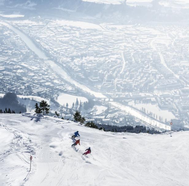 Zwei Skifahrer fahren einen schneebedeckten Hang hinab, im Hintergrund eine verschneite Stadt mit Gebäuden und Straßen.