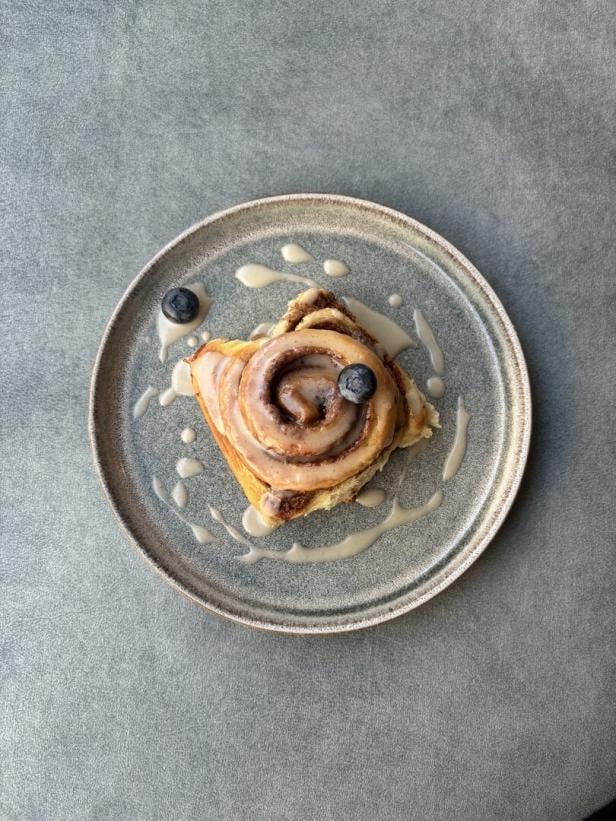 Zimtrolle mit Zuckerguss und zwei Blaubeeren auf grauem Teller, grauer Untergrund.