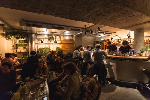 Innenraum einer Bar mit mehreren Gästen an Tischen und an der Theke, warme Beleuchtung und sichtbare Pflanzen an der Wand.