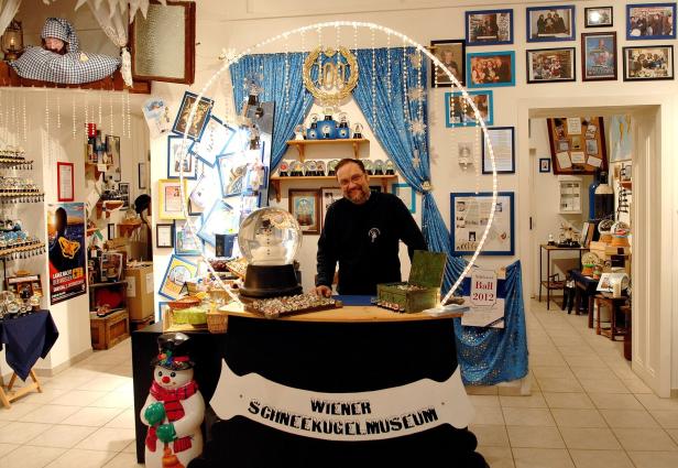 Ein Mann steht hinter einem Tresen im Wiener Schneekugelmuseum, umgeben von Schneekugeln und winterlicher Dekoration.