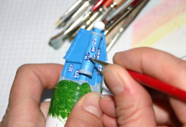 Eine Person bemalt ein kleines, blaues Modellhaus mit einem feinen Pinsel, im Hintergrund liegen weitere Pinsel.