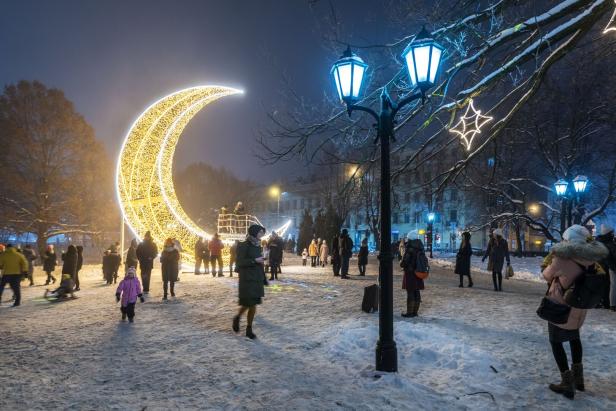 Menschen spazieren in einem verschneiten Park mit einer Lichtskulptur in Form einer Mondsichel und Straßenlaternen.