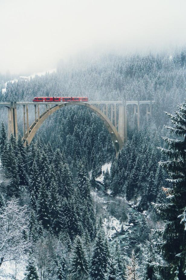 Ein roter Zug fährt in der Schweiz über ein steinernes Viadukt in einer verschneiten Waldlandschaft.