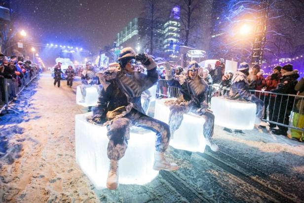 Tänzer in Winterkostümen sitzen auf leuchtenden Eisblöcken bei einer nächtlichen Parade im Schnee.