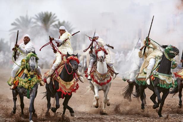 Mehrere Reiter in traditioneller Kleidung reiten auf geschmückten Pferden und halten Gewehre in die Luft, Staub wirbelt auf.