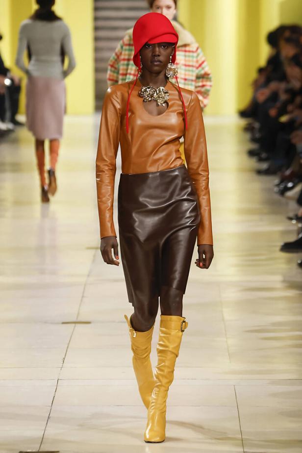 Eine Frau am Laufsteg mit einem braunen Rock, einer roten Haube und gelben Stiefeln.