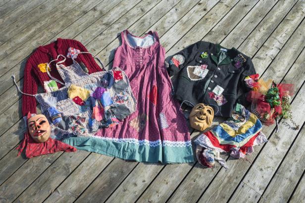 Mit bunten Flicken verzierte Kleidung und zwei Holzmasken liegen auf Holzdielen nebeneinander.