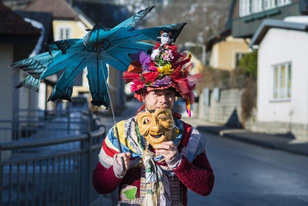 Eine Person mit bunter Verkleidung, auffälligem Hut, zerzaubertem Schirm und Holzmaske steht auf einer Straße.