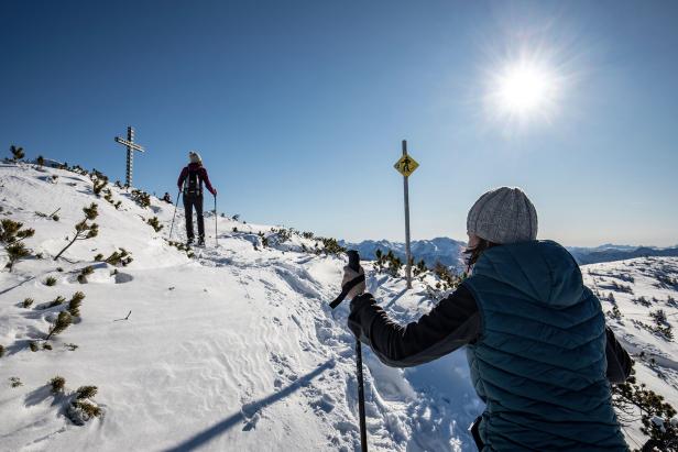 Zwei Wanderer steigen bei Sonnenschein mit Stöcken einen verschneiten Berghang zu einem Gipfelkreuz hinauf.