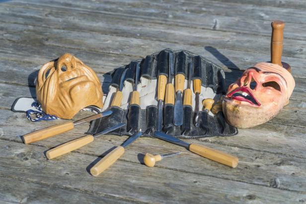 Zwei geschnitzte Masken und verschiedene Holzschnitzwerkzeuge liegen auf einem Holztisch.