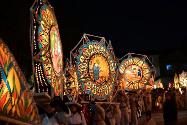Menschen tragen große, bunte, sternförmige Laternen mit gemalten Motiven bei einem nächtlichen Umzug.