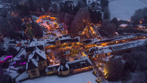 Verschneiter Weihnachtsmarkt mit festlich beleuchteten Ständen und Tannenbäumen in einer historischen Schlossanlage bei Dämmerung.