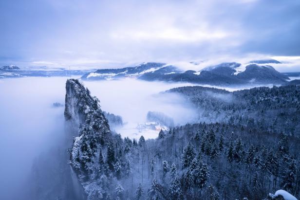 Verschneite Felsen und Tannen ragen aus dichtem Nebel, dahinter Berge und ein Tal mit wenigen Häusern.
