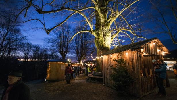 Mehrere Menschen schlendern abends über einen festlich beleuchteten Weihnachtsmarkt mit Holzhütten und Lichterketten.