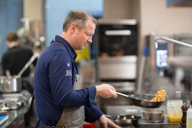 Ein Koch in blauer Kleidung schwenkt gebratene Zutaten in einer Pfanne in einer professionellen Küche.