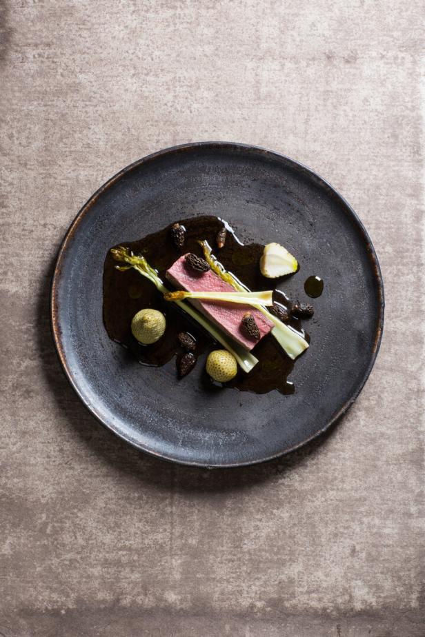 Ein Stück Fleisch mit Spargel, Morcheln, kleinen Kartoffeln und Sauce kunstvoll auf einem dunklen Teller angerichtet.