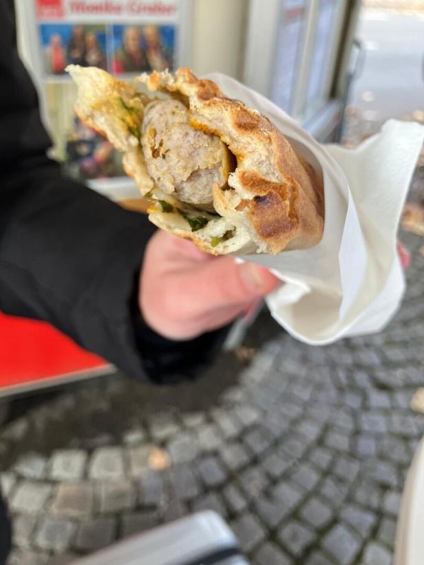 Eine Hand hält ein Brötchen mit einer Bratwurst und etwas Grünem, eingewickelt in eine Serviette.
