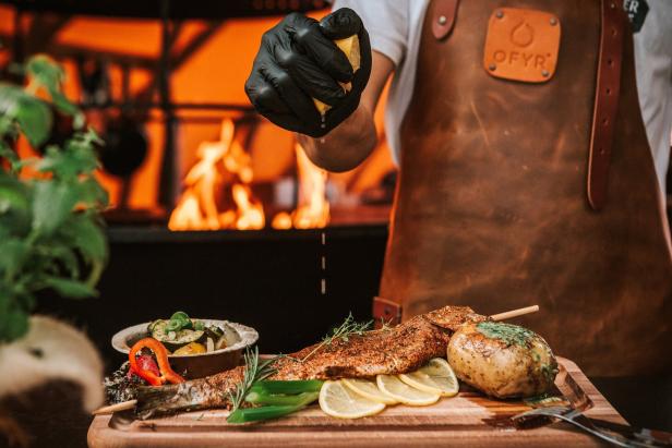 Ein Koch mit Lederschürze presst Zitrone über gegrillten Fisch, Ofenkartoffel und Gemüse auf einem Holzbrett.