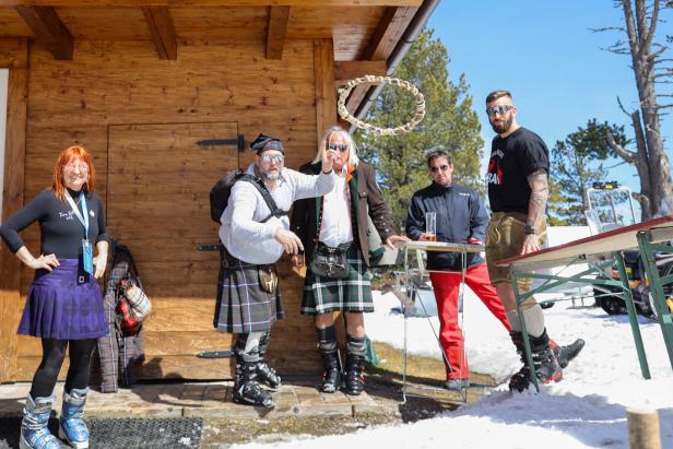 Fünf Personen in Skischuhen und Kilts stehen im Schnee, eine Person wirft einen Ring, andere beobachten oder trinken.