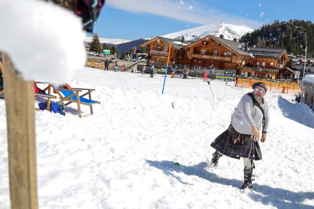 Im Wettbewerb wirft ein Mann im Kilt einen Schneeball vor einer Berghütte im verschneiten Skigebiet.