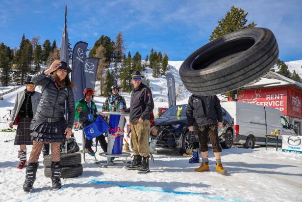 Mehrere Menschen stehen im Schnee, während eine Frau einen Autoreifen wirft und andere zuschauen.