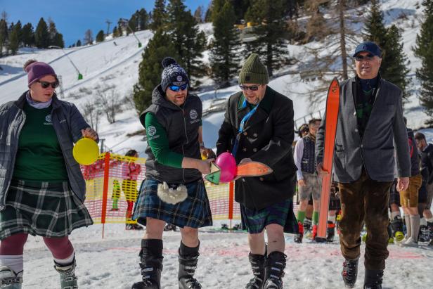 Vier Personen in Skikleidung und Kilts stehen im Schnee, zwei halten Skier und bunte Luftballons.