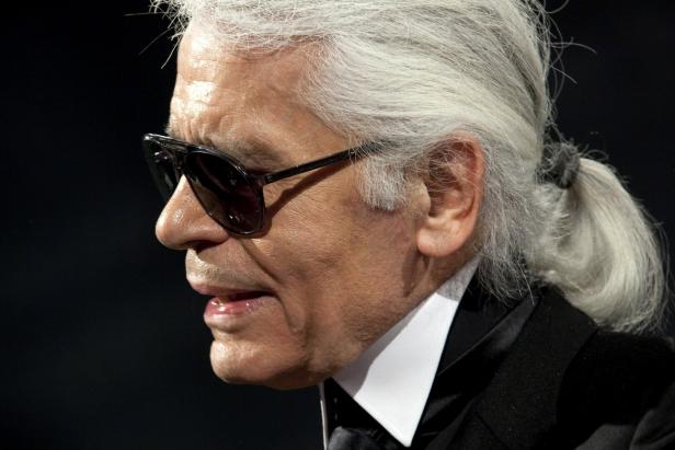 Karl Lagerfeld mit Sonnenbrille und Pferdeschwanz vor dunklem Hintergrund.