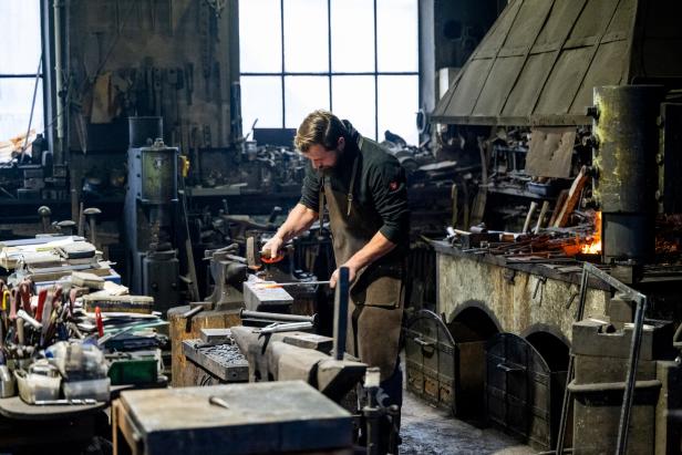 Ein Schmied bearbeitet glühendes Metall auf einem Amboss in einer Werkstatt mit vielen Werkzeugen und einer Feuerstelle.