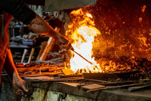 Eine Person hält ein Metallstück ins offene Schmiedefeuer, Funken fliegen und Werkzeuge liegen auf dem Tisch.