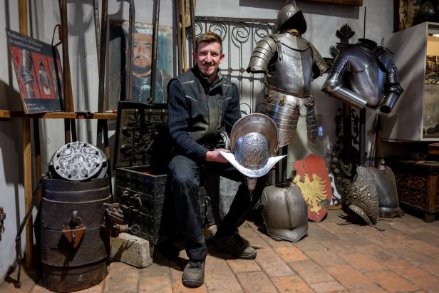 Ein Mann sitzt in einer Werkstatt und hält einen verzierten Helm, umgeben von mittelalterlichen Rüstungen und Ausrüstungsgegenständen.
