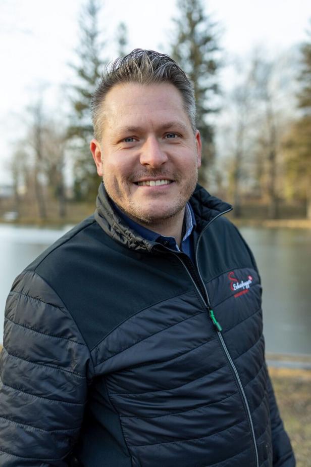 Ein Mann mit dunkler Jacke steht draußen vor einem See und lächelt in die Kamera, im Hintergrund sind Bäume.