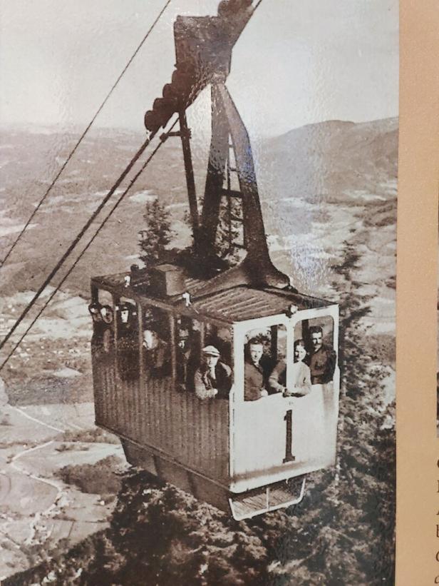 Eine historische Seilbahngondel mit mehreren Menschen schwebt hoch über einer bewaldeten Landschaft.
