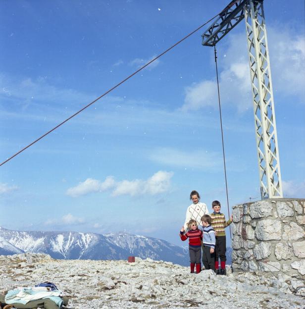 Eine Familie steht auf einem felsigen Gipfel neben einem Skiliftmast vor einer Bergkulisse.
