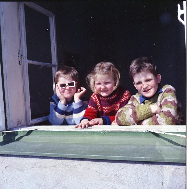 Drei Kinder in bunten Pullovern lehnen lachend an einem offenen Fenster.