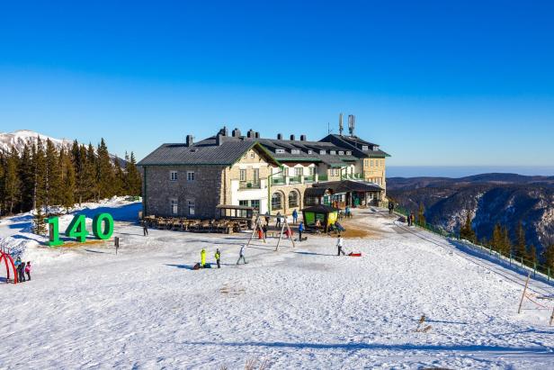 Ein Berghotel mit Gästen liegt auf einer schneebedeckten Anhöhe unter blauem Himmel.
