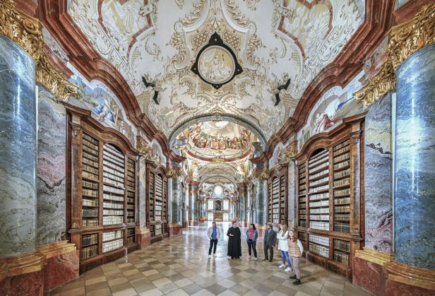 Prunkvoller Bibliothekssaal mit hohen Bücherregalen, verzierten Säulen und einer Gruppe von Menschen in der Mitte.