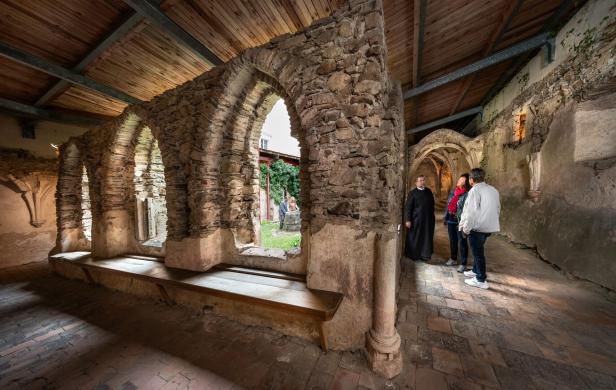 Drei Personen stehen mit einem Mönch in einem alten, überdachten Klostergang aus Steinmauern und Rundbögen.