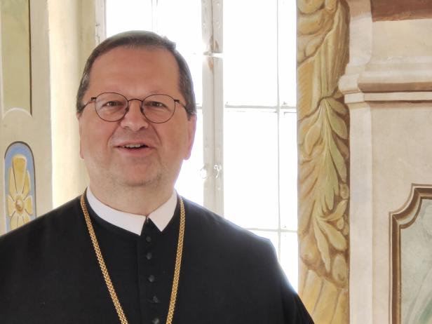 Ein Mann mit Brille und schwarzem Talar mit Goldkette steht vor einem Fenster.