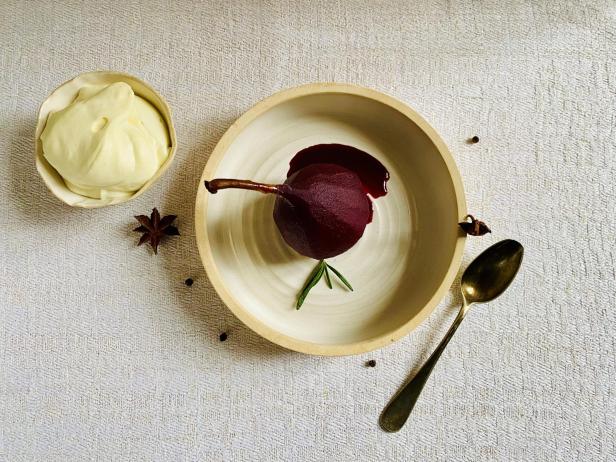 Eine Rotwein-Birne mit Sud und Rosmarin liegt in einer Schale, daneben steht cremige Joghurt-Mousse, umgeben von Sternanis und Pfefferkörnern.
