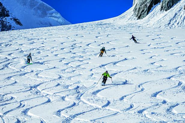 Vier Skifahrer fahren eine verschneite Berghang hinab, der von vielen geschwungenen Spuren durchzogen ist.