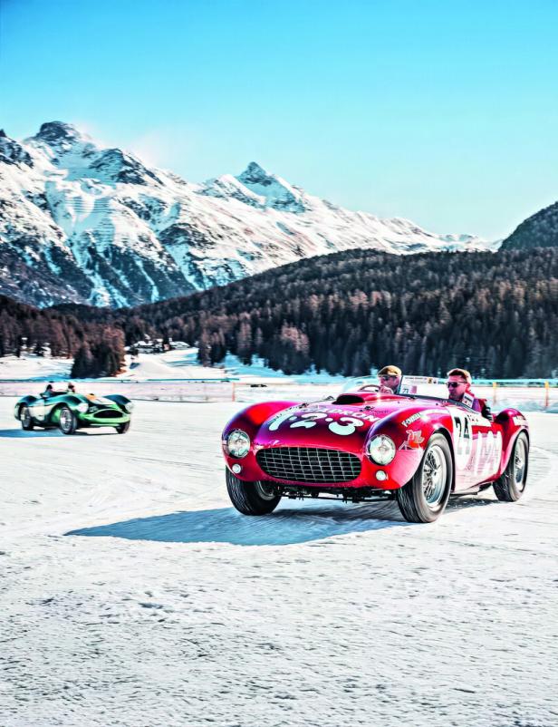 Zwei Oldtimer fahren auf einer schneebedeckten Strecke vor verschneiten Bergen und Wäldern.