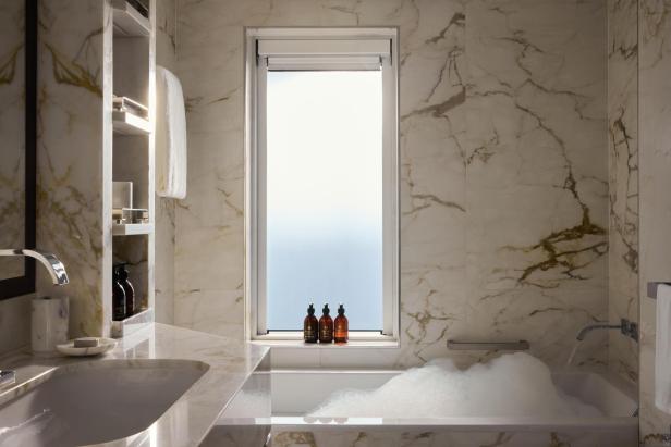 Luxuriöses Badezimmer mit Marmorwänden, Badewanne, Schaum und Pflegeprodukten am Fenster.