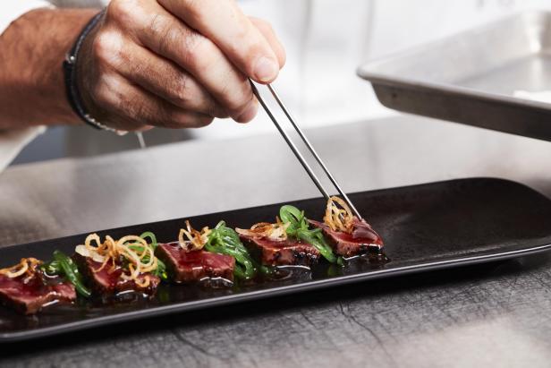 Ein Koch richtet kunstvoll ein Gourmetgericht mit Pinzette auf einem schwarzen Teller an.